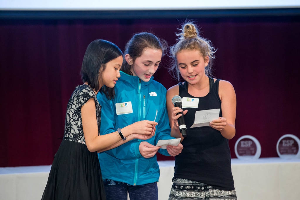 Students reading from notecards into a microphone on stage